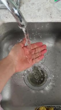 Mutfak musluğundan gelen temiz bir su akıntısının altında durulanan bir elin görüntüsü. Ağır çekimde yakalanan görüntü, akan suyun ferahlatıcı ve arındırıcı hissini vurgular.