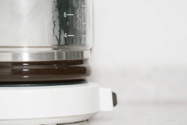 Sürahinin içinden görülebilen taze demlenmiş kahve yapan beyaz damlalı bir kahve makinesi. Buhar yoğunlaşması ve ölçüm işaretleri demleme sürecini vurguluyor. Geniş kopya alanına sahip minimalist beyaz arkaplana karşı ayarla