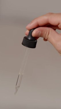 Bir el damlatıcıyı sıkarak temiz bir sıvı damlası bırakıyor, muhtemelen bir cilt serumu ya da nötr gri arka plana karşı gerekli yağ.
