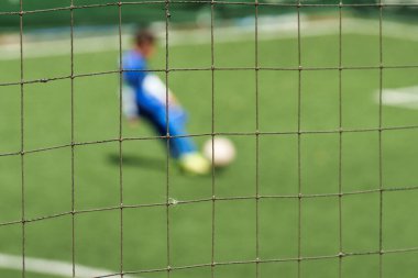 Mavi üniformalı bir oyuncunun yeşil bir yapay çim sahasında topa tekme atmak üzere olduğunu gösteren bulanık bir arka planı olan net bir futbol kalesi.