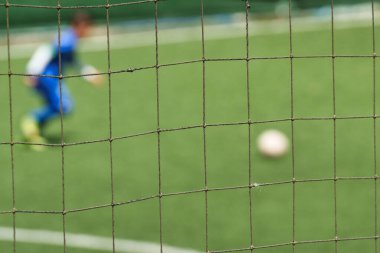 Mavi üniformalı bir oyuncunun yeşil bir yapay çim sahasında topa tekme atmak üzere olduğunu gösteren bulanık bir arka planı olan net bir futbol kalesi.