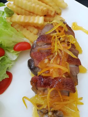Juicy duck confit with orange sauce on white plate. Roasted duck slices topped with orange sauce with grated orange peel, served with French fires and side salad