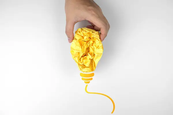 创意和灵感概念 人类的手拿起了在白色背景上隔离的黄色灯泡形状的皱巴巴的彩纸 免版税图库照片