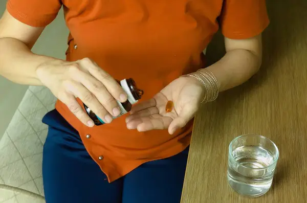 Asian woman holding vitamin capsules in her fingertips, taking vitamins in mouth for treatment, woman taking supplements with water at home to maintain health. Food supplements, vitamins.