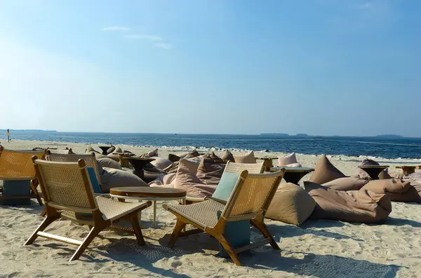 Boş tropikal plaj arka planında sahil kenarındaki bir kafede dinlenme tesisleri olarak masa sandalyeleri ve yastıklar var. Gökyüzü ve beyaz kumla gökyüzü