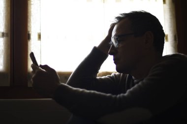 Pencerenin kenarında oturan ve cep telefonu kullanan düşünceli bir adam.