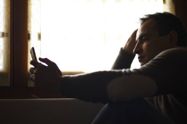 Pencerenin kenarında oturan ve cep telefonu kullanan düşünceli bir adam.