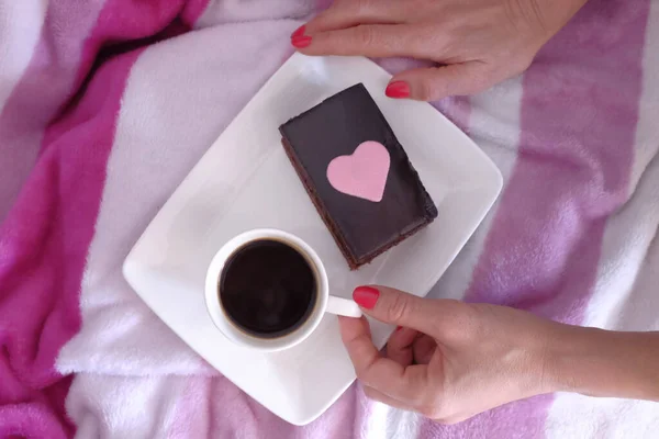 on pink blankets female hands on a tray with cup of coffee accompanied by a chocolate cake with a drawn heart