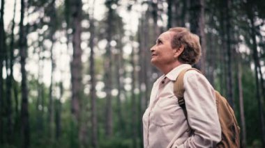 Sırt çantalı yaşlı beyaz bir kadın parkta yürürken temiz havanın tadını çıkarıyor..