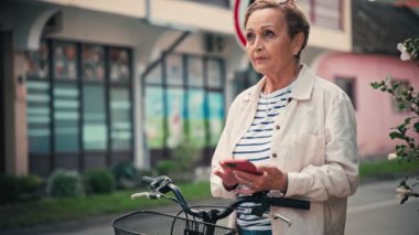 Şehrin göbeğinde bisikletin yanında dikilirken akıllı telefonunu kullanan yaşlı beyaz bir kadının portresi..