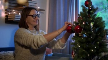 Yavaş çekim sinematik çekim. Genç, beyaz bir kadın yılbaşı için hazırlanırken bir Noel ağacı süslüyor..