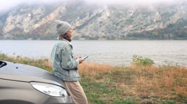 Kafkasyalı genç bir kadın akıllı telefonunu kullanıyor ve araba kaputuna yaslanırken güzel bir dağ manzarasının tadını çıkarıyor..