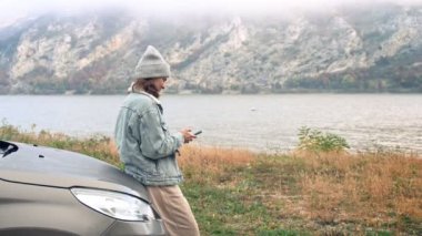 Kafkasyalı genç bir kadın akıllı telefonuyla fotoğraf çekiyor ve araba kaputuna yaslanırken dağların güzel manzarasının tadını çıkarıyor..