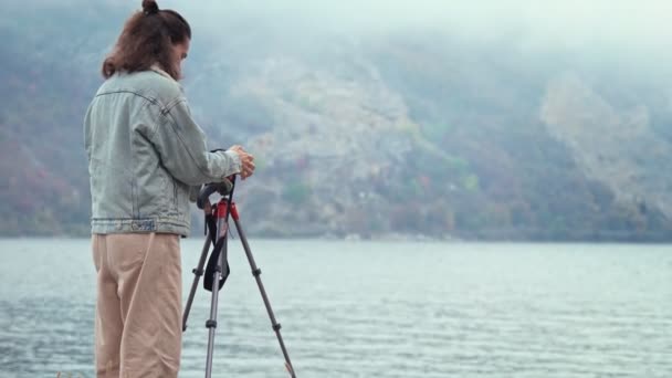 仕事中の若い白人女性プロの写真家 三脚の上にカメラの横に立って 山々の美しい景色を楽しむ人 — ストック動画