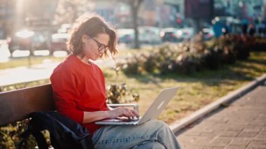 A young caucasian woman wearing glasses typing on the laptop while sitting on the bench outdoors