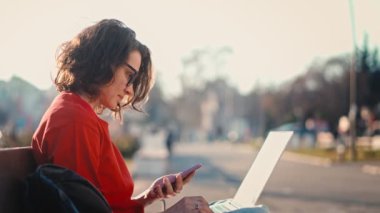 Young caucasian woman in glasses working or studying using a laptop and smartphone while sitting on a bench outdoors