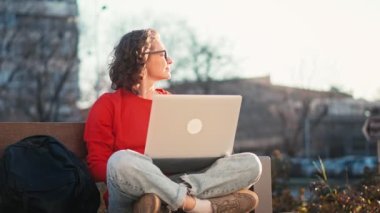 A young caucasian woman wearing glasses typing on the laptop while sitting on the bench outdoors