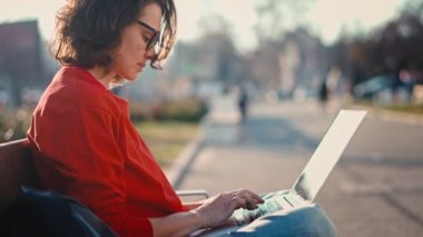 A young caucasian woman wearing glasses typing on the laptop while sitting on the bench outdoors