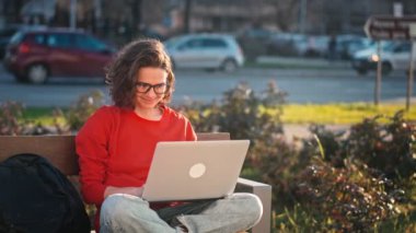 A young caucasian woman wearing glasses typing on the laptop while sitting on the bench outdoors