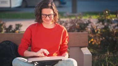 A young caucasian woman in glasses opens her laptop and starts working while sitting on a bench in the street