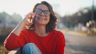 Portrait of a young caucasian cheerful woman talking on the phone while sitting on a bench in the street