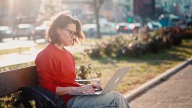 A young caucasian woman wearing glasses typing on the laptop while sitting on the bench outdoors
