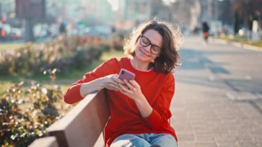 A young caucasian cheerful woman in glasses using her smartphone while sitting on a bench in the street
