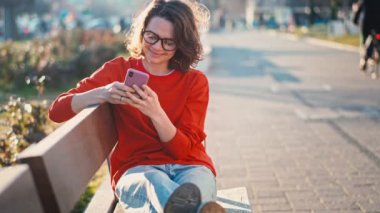 A young caucasian cheerful woman in glasses using her smartphone while sitting on a bench in the street