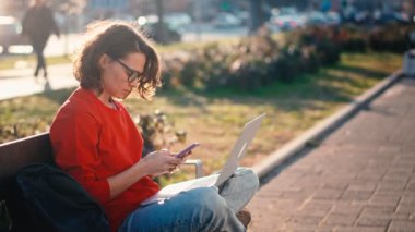 Young caucasian woman in glasses working or studying using a laptop and smartphone while sitting on a bench outdoors