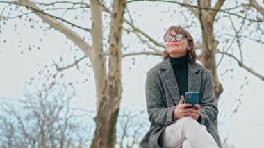 A slow-motion shot of a caucasian woman wearing glasses using her phone while sitting on the bench outdoors