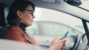 A slow-motion shot of a caucasian businesswoman in glasses using her smartphone while sitting in the drivers seat of a parked car.