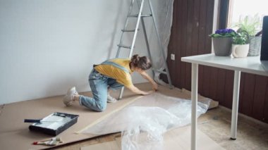 A young caucasian woman is preparing a room for painting. A female fixing the film with masking tape while standing on a stepladder.