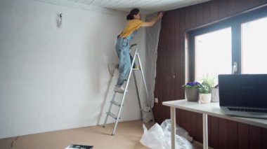 A young caucasian woman is preparing a room for painting. A female fixing the film with masking tape while standing on a stepladder.