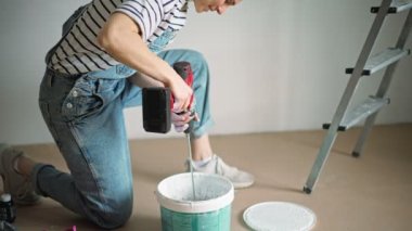 A young caucasian woman mixing paint in a bucket using a cordless drill with a mixing nozzle.