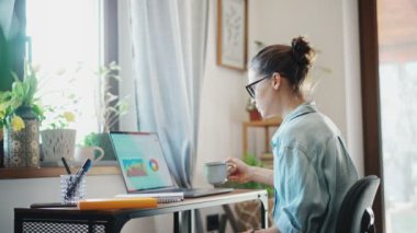 A young caucasian woman using a laptop and making notes in a notebook while sitting on a computer chair in a home office.