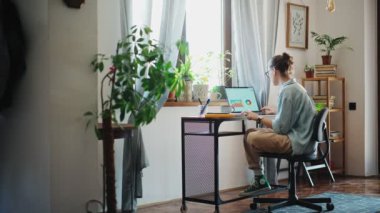 A young caucasian woman using a laptop and making notes in a notebook while sitting on a computer chair in a home office.