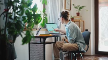 A young caucasian woman using a laptop and making notes in a notebook while sitting on a computer chair in a home office.