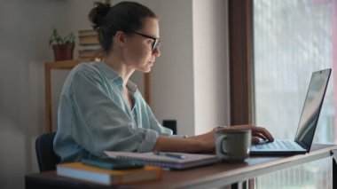 A young caucasian woman wearing glasses typing on a laptop while working from home.