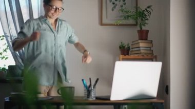 Genç, beyaz bir kadın ev ofisinde çalışırken laptopunun önünde oynaşıp dans ediyor..