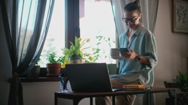 A young adult caucasian woman drinking coffee and using a smartphone while working from home.