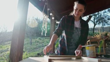 A young caucasian woman using a brush to varnish a wooden board while standing at sunny terrace of her country house.