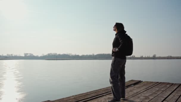 Задумчивая Женщина Рюкзаком Стоящая Пирсе Замёрзшего Озера Смотрящая Воду Солнце — стоковое видео