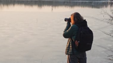 Yavaş çekim sinematik çekim. Günbatımında gölün kenarında duran profesyonel bir fotoğrafçı kadın manzaranın fotoğrafını çekiyor.