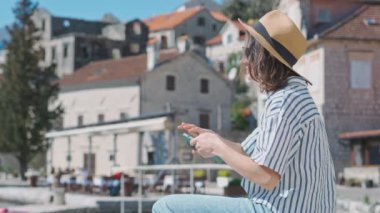 Eski bir Avrupa sahil kasabasının arka planında otururken şapkalı genç bir beyaz kadın telefonuyla fotoğraf çekiyor.