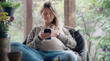 Hamile, orta yaşlı bir kadın sıcak bir sonbahar gününde bir kulübenin terasında otururken akıllı telefon kullanıyor.