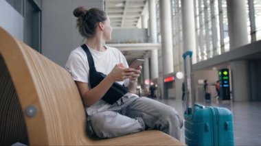 Elinde telefon olan genç bir bayan gezgin havaalanı bekleme salonunda bir bankta oturuyor.