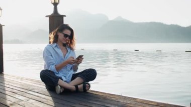 Kafkasyalı genç bir kadın iskelede otururken rahatlar ve akıllı telefonunu kullanır.
