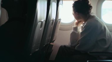Uçuş sırasında uçak koltuğunda oturan genç bir kadın pencereden dışarı bakar.