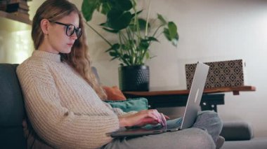 Genç ve güzel bir kadın bir kış akşamı rahat bir oturma odasında kanepede otururken dizüstü bilgisayarda çalışıyor.