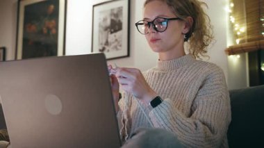 Genç ve güzel bir kadın, kış akşamları rahat bir oturma odasında kanepede otururken internette örgü örmeyi öğreniyor.